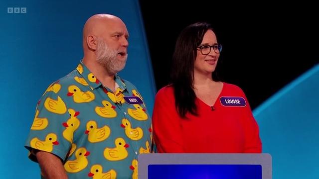 A balding white man in his fifties wears a short-sleeved shirt, it's water blue with lots of pictures of rubber ducks floating about. He stands next to a long-haired woman who wears a bright red top.