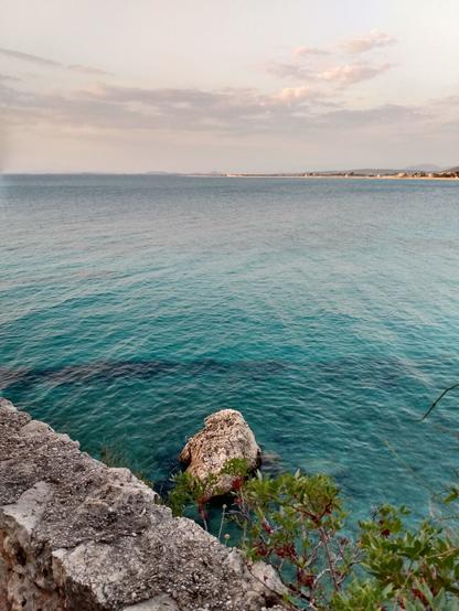 An old grey stone wall can be seen in the bottom left corner of the picture. Below the edge is a small rock in the water. Behind it, the clear turquoise-coloured sea stretches towards the horizon. A headland juts into the picture from the right in the distance under a hazy sky.