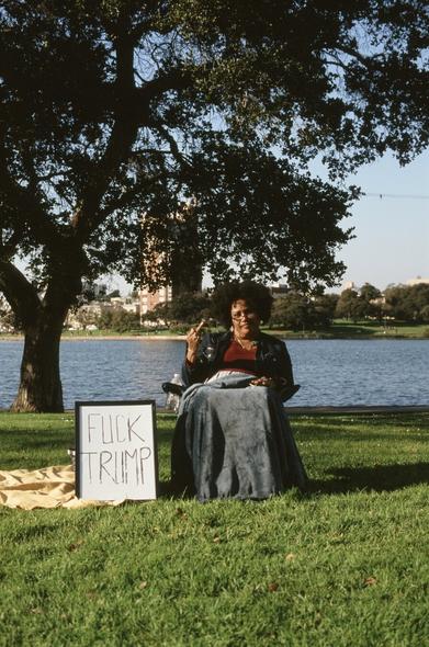 A black woman is seated with a grey-blue blanket over her legs in a maroon blouse and black jacket. Her hair is curly behind a band and she is wearing glasses while raising a middle right finger. Next to her on a white board is a whiteboard that says ‘fuck Trump’ on a blanket. She is seated on the green lawn near a tree with lake Merritt and the edge of downtown Oakland in the distance.