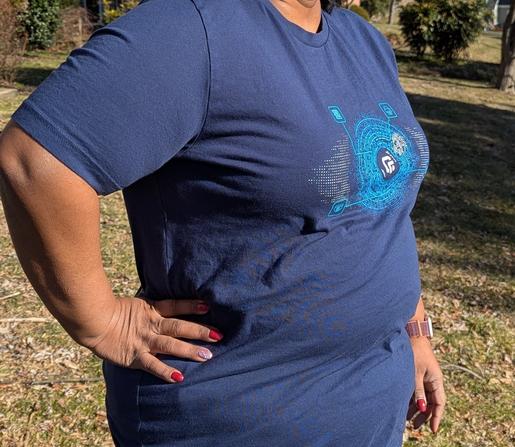 Photograph of a woman standing in bright sunlight obliquely facing camera right, pictured neck down and waist up with her right arm resting on her hip. She wears a dark blue t-shirt with a runZero logo surrounded by hacker-cool symbolism on the chest.
Behind are grass, leaves, twigs, bushes and trees of the type which may be found on a front lawn.