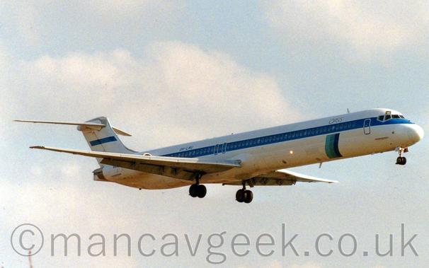Side view of a white, twin engined jet airliner with the engines mounted on the sides of the rear fuselage, flying from left to right at a very low altitude, with it's undercarriage lowered and flaps extended from behind the wings, suggesting it is about to land.
The plane has a thick blue stripe running along the body, covering the passenger cabin windows, and small black "Oasis" titles on the upper forward fuselage.
Under the "Oasis" titles, a vertical bar in 3 shades of blue extends down towards the plane's belly.
Pale blue sky with hazy white cloud fill the rest of the frame.