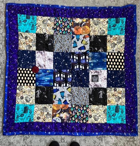 A square quilt made from 36 squares of printed fabric arranged in a 6x6 square with a purple border and dark blue binding; the fabrics are all in prints referring to Doctor Who, space, or time. The dominent colors are dark blue, turquoise, grey, and pale gold.