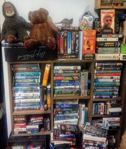 A very cluttered bookshelf full of VHS tapes facing all directions, with a teddy bear and a stuffed Bigfoot from "Harry and the Hendersons" sitting on top.