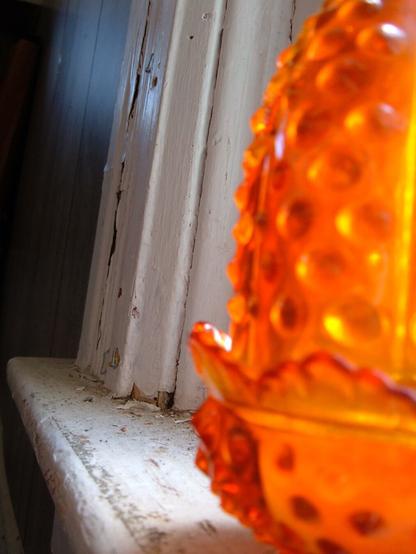 A cropped closeup of a vintage orange hobnail glass candle holder in the foreground, sitting on a windowsill with chipped white paint in focus behind it.