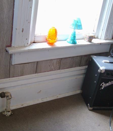 A shot looking down at the orange glass candle holder and a turquoise lamp candle holder, sitting on the windowsill with bright light coming in through the window. There is a black Fender amplifier on the gray floor beneath the window.