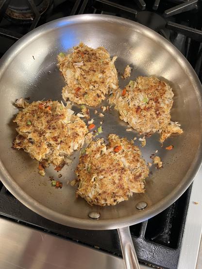4 toasty brown crab cakes, sautéeing in a silver frying pan, bits of bell pepper and celery