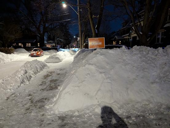 Snowbank next to sidewalk in residential neighborhood at night with an orange Ontario NDP election sign on top