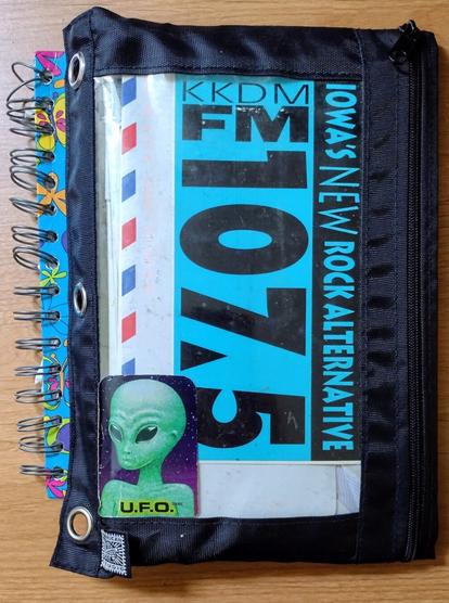 A black zippered pencil case with a clear window showing the contents inside. A turquoise and black bumper sticker for Iowa's 107.5 radio station fills the frame. The red, white, and blue border of a U.S. Army Airmail envelope can also be seen. A sticker with a green alien and the letters U.F.O. is stuck on the outside.