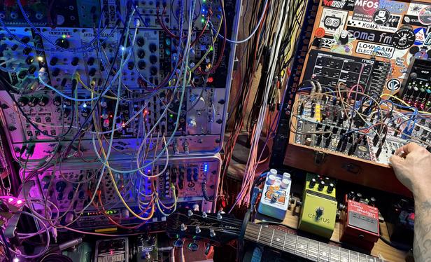 Wide view across dozens of synth modules in a busy patch filled with different colored cables, illuminated from different angles by different shades and colors of light. It is a complex jumble of many things.