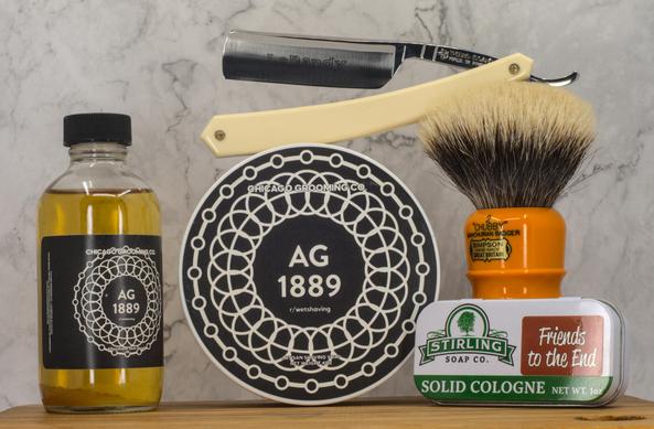 (Le Chat Mistral description)
The image displays five grooming items arranged on a wooden surface against a marbled grey background. From left to right: a small bottle labelled "Chicago Grooming Co. AG 1889" containing a yellow liquid. Next, a round tin of "AG 1889" shaving soap with a black and white design. Above the tin, a Thiers Issard Le Dandy straight razor with white scales. Beside it, a shaving brush with an orange handle and light bristles, labelled "Chubby." Lastly, a small tin of "Stirling Soap Co. Friends to the End" solid cologne. The items are neatly presented, suggesting a shaving or grooming setup.