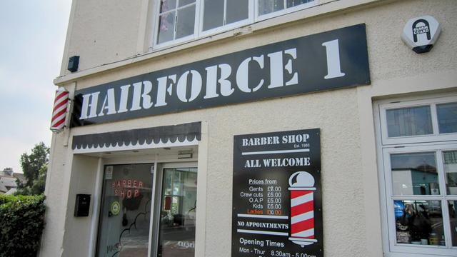 Exterior of Hairforce 1 barber shop, a beige building with a black sign and striped barber pole, located in Penzance, UK. The sign lists services and prices.