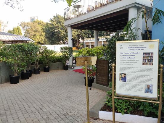 A large poster outside of the entrance to the venerable Bangalore Club library. There are lots of potted plants, columns at the entrance. Members only.