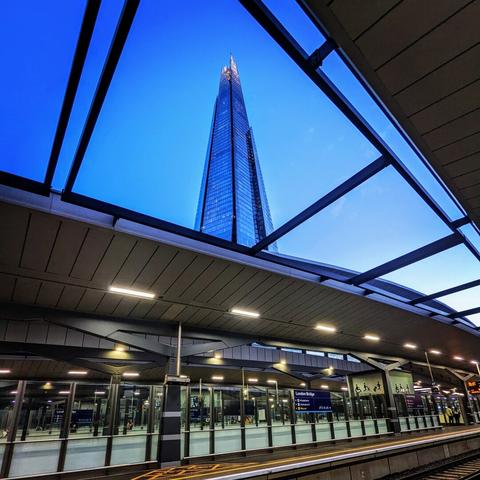 View from London Bridge Station platform, looking up at The Shard skyscraper illuminated against a deep blue evening sky. The station's glass and metal roof structure is visible, along with platform markings, and signage (including "London Bridge" and restroom symbols).