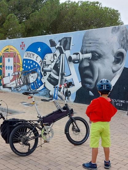 Criança ao lado de uma bicicleta dobravel VELLO cinza escura a olhar para um mural onde está Gago Coutinho e as bandeiras de Portugal e Brasil