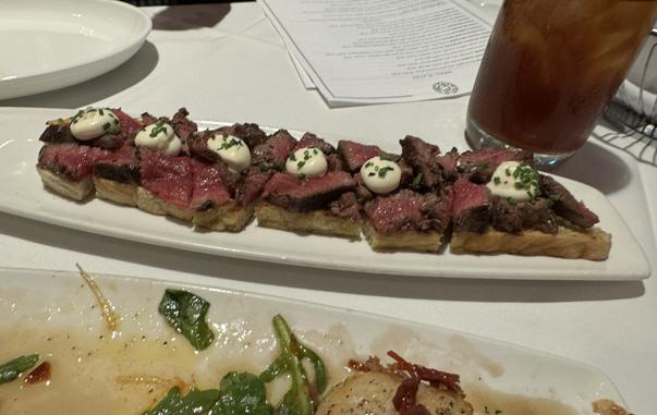 A platter of rare steak on toasted bread, topped with dollops of creamy sauce and fresh chives. In the background, there's an empty plate with remnants of a salad. A glass of iced tea is also visible.