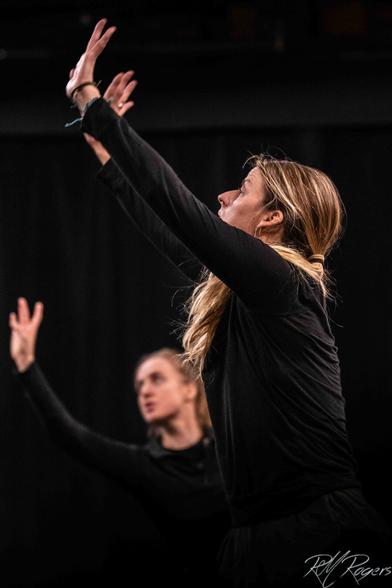 Two women in black clothing, against a black background, perform a modern dance routine with arms and hands outstretched.