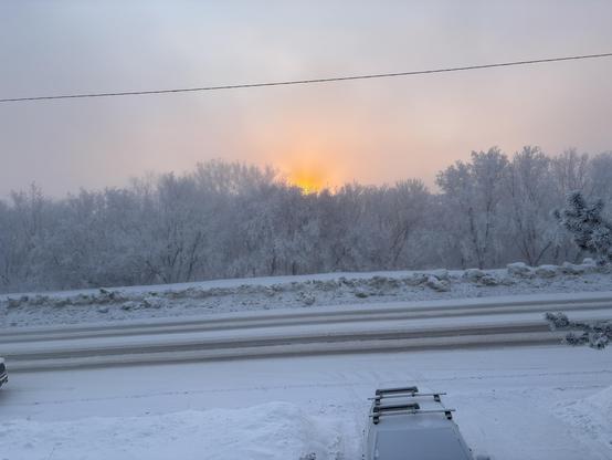 A snow covered road.. Behind are trees covered in hoarfrost. Clouds of steam are rising from the river. The rising Sun glows Orange