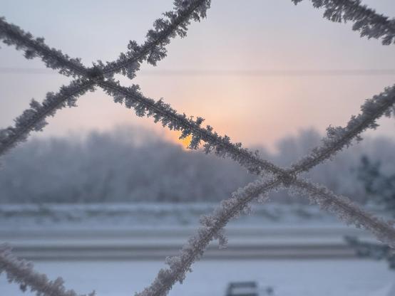 The same landscape as in the other picture is blurred in the background. In three foreground, there is a net with a diamond structure. The threads are covered with thick ice crystal formations