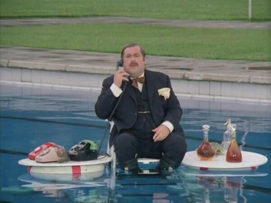 A photo of a heavy man with a mustache, wearing a suit. He is sitting on a floating chair in a swimming pool, talking on a telephone. His legs are dangling in the water. The phone is sitting with two others on a life preserver ring to the man's right. To his left is another life preserver ring, on top of which sits a tray with a spritzer bottle and some decanters, presumably filled with types of liquor.
