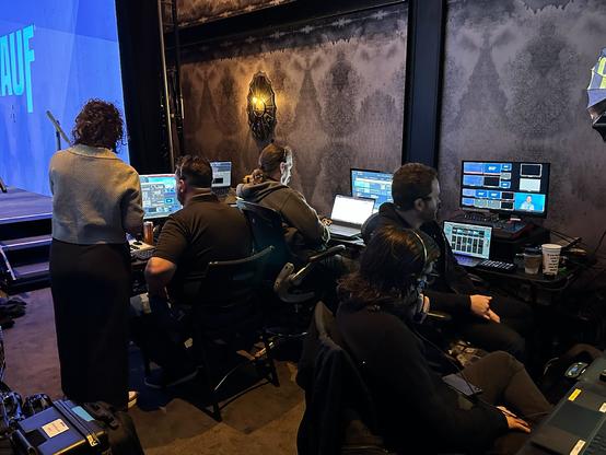 Backstage area with multiple computer workstations, monitors, cables, and equipment on tables. Five people are in the work area.