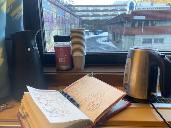 Picture of a open book in the window. Behind the book there is a water boiler, a coffemug, instant coffe and papercups.