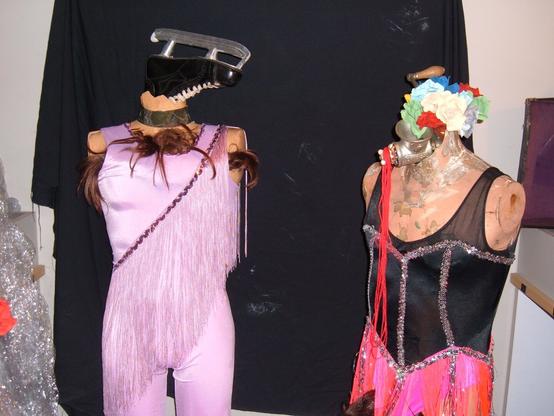 An assemblage of objects displayed against a black curtain: Two armless, headless mannequins; one wearing a pink leotard with long fringe and an ice skate in place of its head, and another wearing a black and silver sequin costume with neon pink fringe, with fake flowers in place of its head.