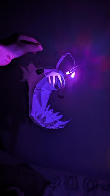 a low poly paper model of an anglerfish light with an RGB LED. The LED lights up purple tinting the otherwise dark room and highlighting the huge mouth and the sharp teeth of the anglerfish