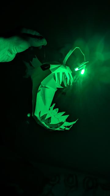 a low poly paper model of an anglerfish light with an RGB LED. The LED lights up green, tinting the otherwise dark room and highlighting the huge mouth and the sharp teeth of the anglerfish