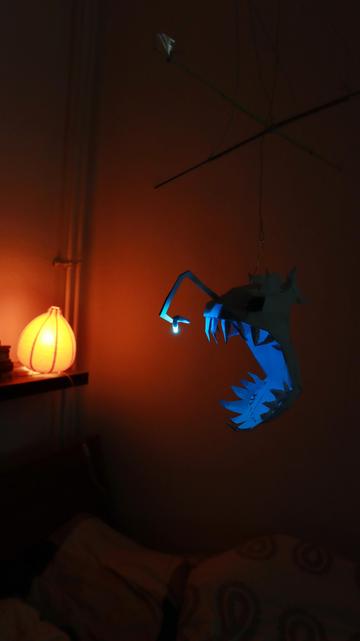 a low poly paper model of an anglerfish light with an RGB LED. The LED lights up blue tinting the highlighting the hughe mouth and the sharp teeth of the anglerfish. the background shows a bed at wall. The room is dark only light by a red lamp and the anglerfish