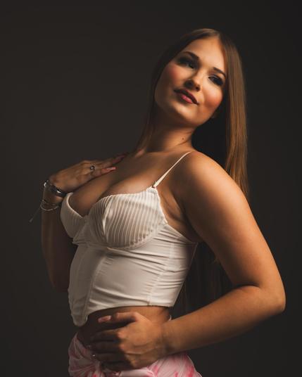 The image features a person posing in front of a dark background. They are wearing a white corset-style top and have long hair. The lighting focuses on the person, highlighting their facial features and attire.