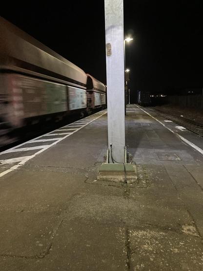 In der Mitte steht ein Betonpfeiler. Rechts und links Bahnsteig und Gleise. Rechts fährt ein Güterzug vorbei. Es ist dunkel. Der Bahnsteig ist durch mit künstlichem Licht beleuchtet.