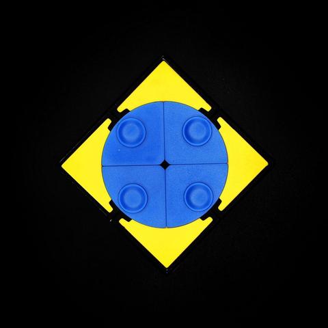 Vista cenital de un cubo sobre fondo negro.
El cubo está girado 45º y solo podemos ver la cara superior amarilla didivida en cuatro piezas (es un 2x2x2). Sin embargo, justo en el medio hay un círculo de color azul dividido, a su vez, en cuatro piezas. Cada una de esas piezas tiene un relieve circular.
La fotografía es muy MUY simétrica.