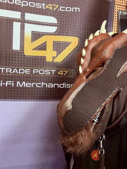 A person wearing a dragon mask poses in front of a backdrop displaying the logo "Trade Post 47." He holds something furry between his teeth.