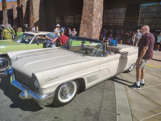 A 1960 Lincoln two-door convertible with its classic massive proportions and gingerbread, all of which would be dropped for the 1961 model year. This example is off-white with a golden tan leather interior.  The picture is from eye height on the driver's side, a front three-quarter view.