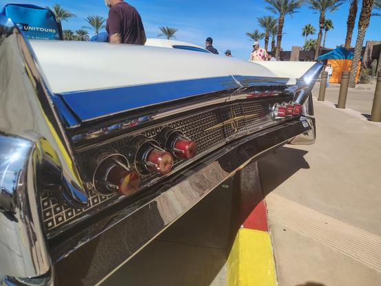 The elaborate rear chrome molding, fins, and taillights of a 1960 Lincoln convertible.  The taillight surround is a series of 1 inch matte black squares each with a chrome center dot and each surrounded by chrome.  It extends the width of the car, and features a gold-tone Lincoln emblem in the center. A deep chrome band extends from the bumper up the sides of the car and its tail fins, the across the trailing edge of the trunk.