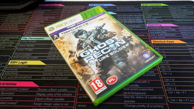 Zdjęcie przedstawia pudełko z grą „Tom Clancy's Ghost Recon: Future Soldier” na konsolę Xbox 360. Na okładce widnieje dynamiczna grafika – uzbrojony żołnierz w futurystycznym pancerzu i hełmie taktycznym znajduje się na tle pola bitwy, z unoszącym się pyłem i eksplozjami. W lewym dolnym rogu widoczne jest oznaczenie wiekowe „18+” oraz symbol „PL”, a w prawym dolnym rogu logo Ubisoft. Pudełko ma charakterystyczny zielony kolor, typowy dla gier na Xbox 360. W tle zdjęcia znajduje się kolorowa podkładka z tabelą komend systemu Linux.