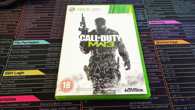 Zdjęcie przedstawia pudełko z grą „Call of Duty: Modern Warfare 3” na konsolę Xbox 360. Na okładce widnieje sylwetka żołnierza w bojowym stroju, trzymającego karabin, na tle rozmytego, białego dymu i czarnych plam. Tytuł gry jest wyeksponowany w centralnej części okładki, z charakterystycznym zielonym napisem „MW3”. W lewym dolnym rogu znajduje się oznaczenie wiekowe „18+”, a w prawym dolnym logo Activision. Pudełko ma zieloną ramkę, typową dla gier na Xbox 360. W tle zdjęcia widoczna jest kolorowa podkładka z tabelą komend systemu Linux.
