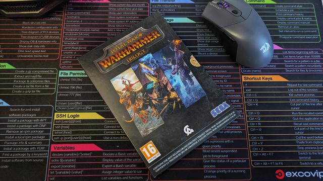 Zdjęcie przedstawia pudełko z grą „Total War: Warhammer Trilogy”. Na okładce widoczne są dynamiczne grafiki z trzech części serii – przedstawiają epickie sceny bitewne z udziałem fantastycznych stworzeń, takich jak smoki, wojownicy chaosu i inne ikoniczne postacie z uniwersum Warhammera. Tytuł gry „Total War: Warhammer Trilogy” jest wyeksponowany złotymi literami na ciemnym tle. W lewym dolnym rogu znajduje się oznaczenie wiekowe „16+”, a w prawym dolnym logotypy SEGA i Creative Assembly. Pudełko leży na kolorowej podkładce z tabelą komend systemu Linux, obok myszki gamingowej Redragon.