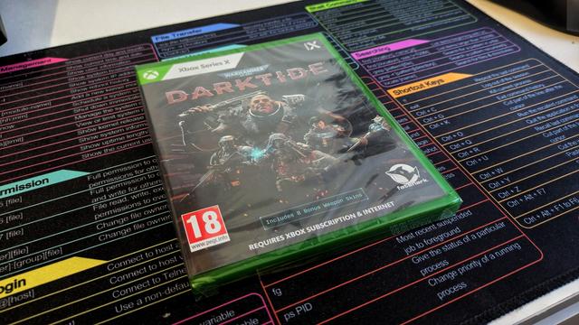 Zdjęcie przedstawia pudełko z grą „Warhammer 40,000: Darktide” na konsolę Xbox Series X. Na okładce widnieje dynamiczna grafika przedstawiająca grupę bohaterów w futurystycznych zbrojach, otoczonych mroczną atmosferą i gotowych do walki. Tytuł gry „Darktide” jest wyeksponowany czerwonymi literami w centralnej części okładki. W lewym dolnym rogu znajduje się oznaczenie wiekowe „18+”, a w prawym dolnym logo Fatshark. Pudełko ma zieloną ramkę, charakterystyczną dla gier na Xbox. W tle zdjęcia znajduje się kolorowa podkładka z tabelą komend systemu Linux.
