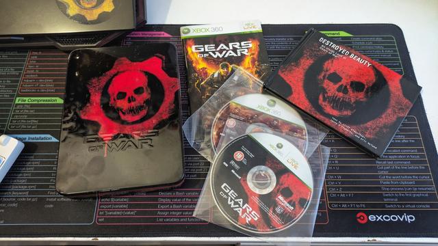 Zdjęcie przedstawia zestaw kolekcjonerski gry „Gears of War” na konsolę Xbox 360. Na pierwszym planie widoczne są: metalowe pudełko z charakterystycznym czerwonym logo czaszki, standardowe pudełko z okładką gry, książeczka „Destroyed Beauty: An Inside Look at Gears of War” oraz dwie płyty – jedna z grą, druga z dodatkowymi materiałami. Wszystkie elementy leżą na kolorowej podkładce z tabelą komend systemu Linux. W tle widać fragment laptopa z naklejką z logo Gears of War oraz fragment klawiatury.