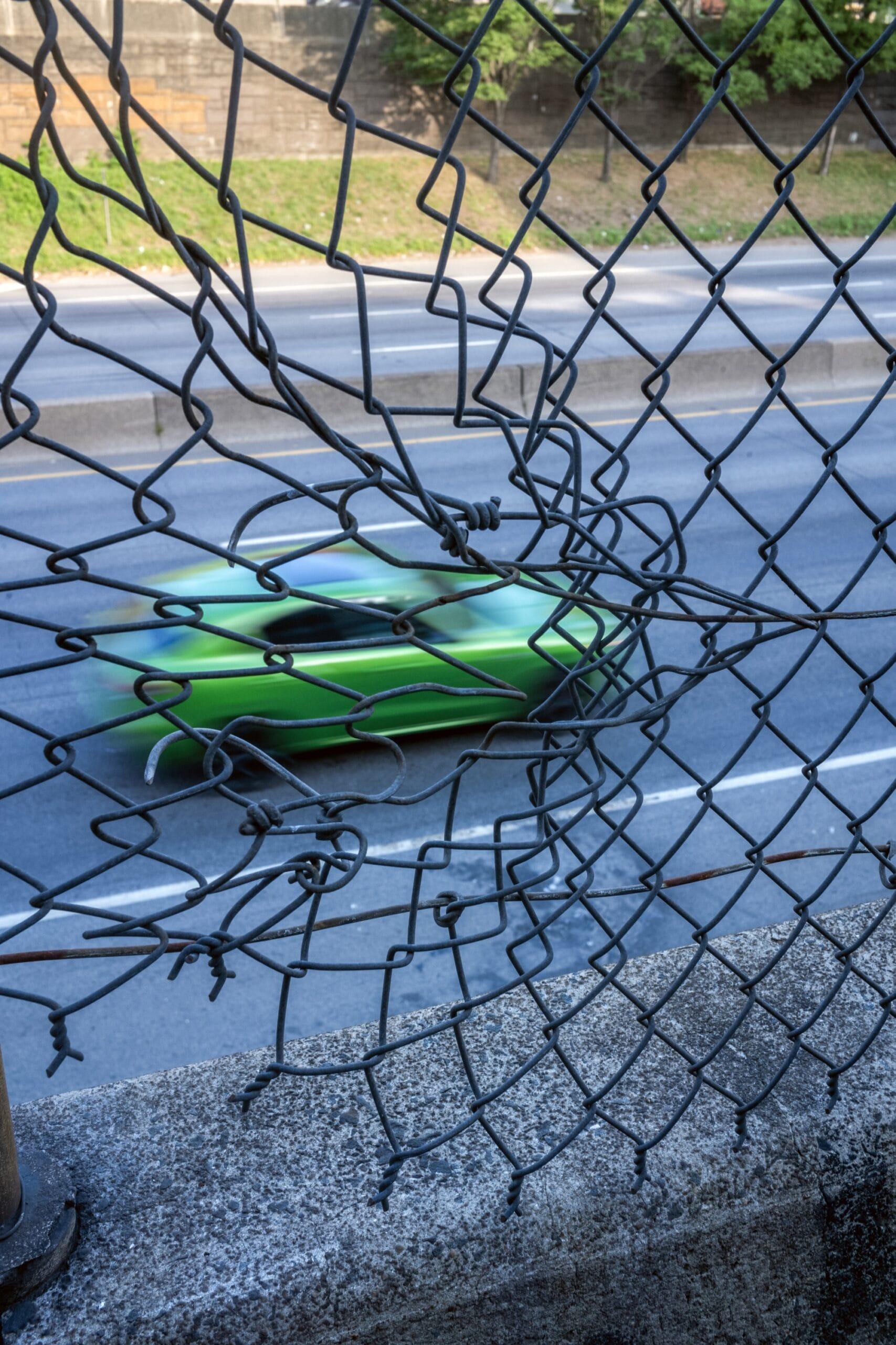 a photograph of a green car speeding along a road, seen through a chainlink fence that appears to be moving with the car