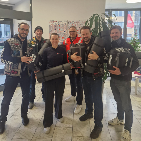 Member des Chapter Frankfurt im EIngangsereichder Johanniter Unfalhilfe Rhein-Ruhr. SIe halten Schlafsäcke und Isomatten, Im Hintergrund sind große Glasfenster, eine Sitzecke und eine große Grünpflanze.