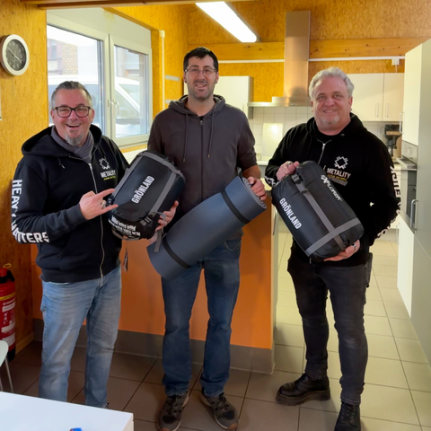 3 Personen mit Schlafsäcken und Isomatten - 2 Metality-Member und der Mitarbeiter der Hilfsorganisation “Treff em Gässje”. Sie befinden sich in einem Gemeinschaftsraum, hinter ihnen befindet sich eine Küche.