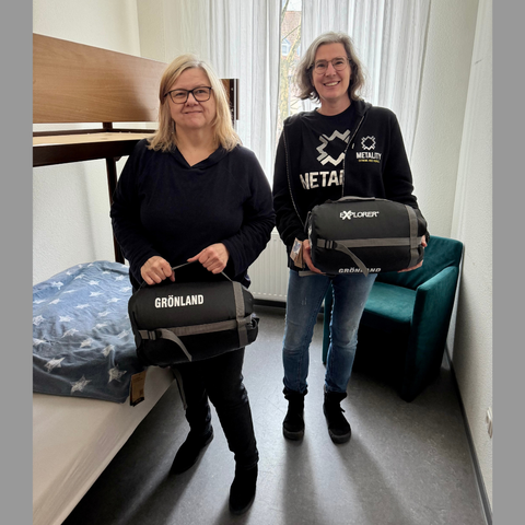 Zwei Frauen haben sich für ein Foto in einem Notunterkunftszimmer der Wohnungslosenhilfe Oase in Saarlouis aufgestellt. Jede der beiden hält einen symbolischen Schlafsack in den Händen. Neben den Personen befindet sich ein Etagenbett und hinter ihnen ein Sessel, der vor einem Fenster aufgestellt ist.