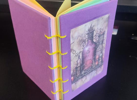 Handmade notebook, hardcover with exposed spine. Binding style: coptic stitch, using yellow thread. Book cover purple. On the front cover there is an image of a purple fantasy style bottle with a light-purple liquid inside.