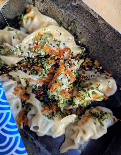 Look into a black food delivery box out of cardboard with dumplings, coated with roasted onions an some green herbs.