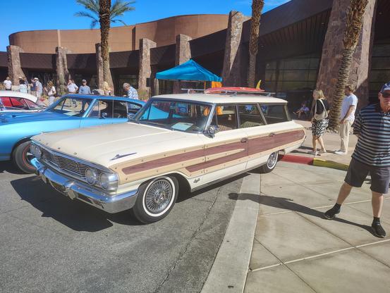 A cream 1964 Ford Country Squire wagon with a tan interior.  Since this is California, there's a surfboard strapped to the luggage rack.  The badge just forward of the driver's door indicates it has the optional 390 engine. 

Just behind it is a 68 GTO (correct me on the year if I'm wrong, but head restraints became required equipment in the US in 1969, and that GTO has none, so I think the model year is correct.)