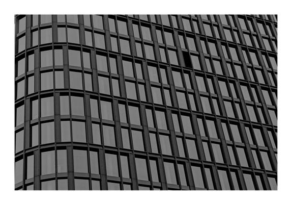 The image shows a black and white photograph of the facade of a modern building. The building has a grid-like pattern created by numerous windows and vertical as well as horizontal structural elements. The windows are arranged in a repetitive manner, contributing to a structured and geometric aesthetic.