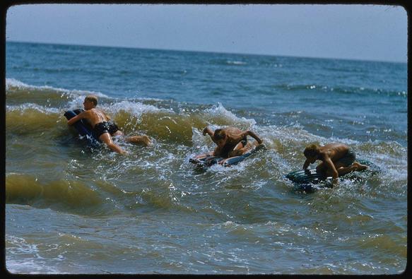 The photograph depicts three young boys engaging in beach activities on a sunny day, as captured by American photographer Toni Frissell. The scene is set along the shoreline of Long Island's scenic beaches during the 1940s era.

In this color transparency image, each boy appears to be using a small floating device or surfboard while riding ocean waves that gently break near the shore. Their expressions and body language suggest they are enjoying themselves as they navigate through the water. The boys' clothing is indicative of mid-20th-century beachwear: swim shorts in darker shades.

The photograph's composition captures the essence of leisurely summer activities during a bygone era, showcasing both playful action and serene relaxation within an idyllic coastal setting. Frissell's work often encapsulates American life experiences with grace and elegance, as seen here through this vibrant snapshot from The Hamptons collection for Sports Illustrated.

Keywords: beach scenes, Long Island, the Hamptons, way of life