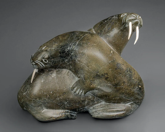 official museum photo of the object, front profile on grey background: Steatite (soapstone, greenish-brown with darker blackish areas and whitish veins throughout) sculpture of two tusked walruses huddled together, with facial features and flipper details incised